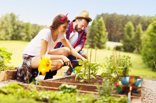 A man and a woman are working together in a garden with a raised wooden bed, tending to young vegetable plants. The woman, wearing a white t-shirt and plaid shorts, has a pink headband, while the man, dressed in a red and blue checkered shirt, hat, and jeans, is using gardening tools. The garden features soil beds filled with green seedlings, bordered by gravel pathways. In the foreground, a small yellow watering can and colorful plant markers are visible. The background reveals lush, green trees and a bright, sunny sky, indicating a warm day. The scene is part of a well-maintained outdoor space, typical of a suburban garden in Gunnersbury, with natural textures of soil, wooden planters, and vibrant foliage that showcase outdoor gardening and lawn care activities associated with professional gardening services such as Lawn Mowing Gunnersbury. The overall setting emphasizes a thriving and healthy garden environment prepared for ongoing planting and maintenance, designed to support local garden enhancement efforts.